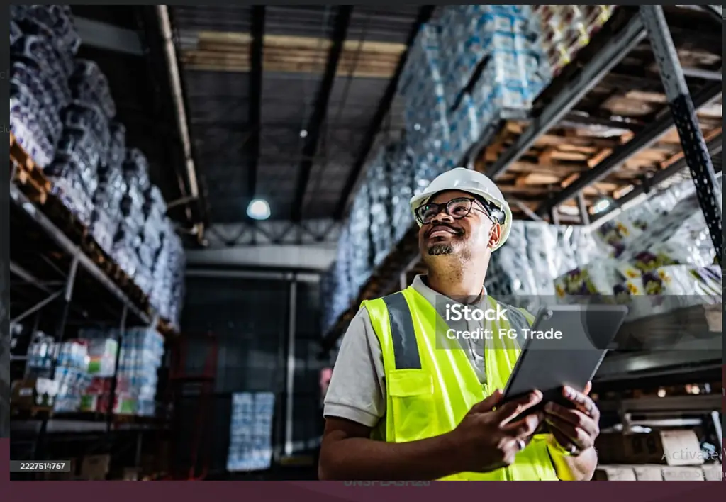 Warehouse supervisor in a safety vest and hard hat reviewing operations on a digital tablet
